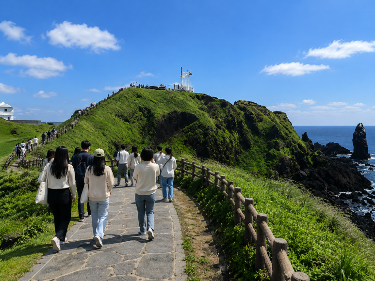 푸른 하늘 아래 초록빛 언덕과 등대가 보이는 성산일출봉 산책로를 걷는 사람들